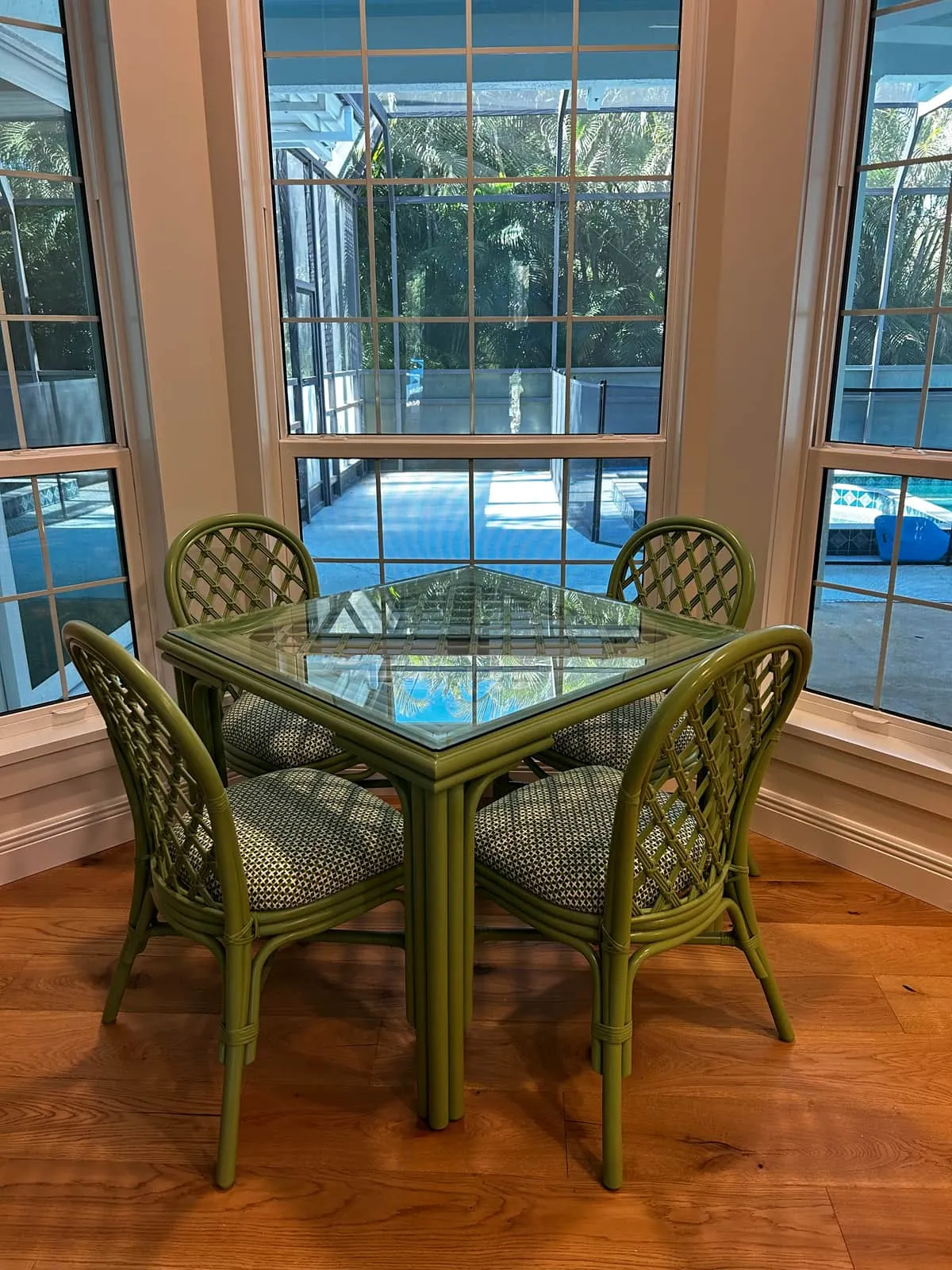 A square glass-topped table and four lattice-back chairs with patterned seat cushions, all painted olive green, standing in a bay-window nook over a wood floor.