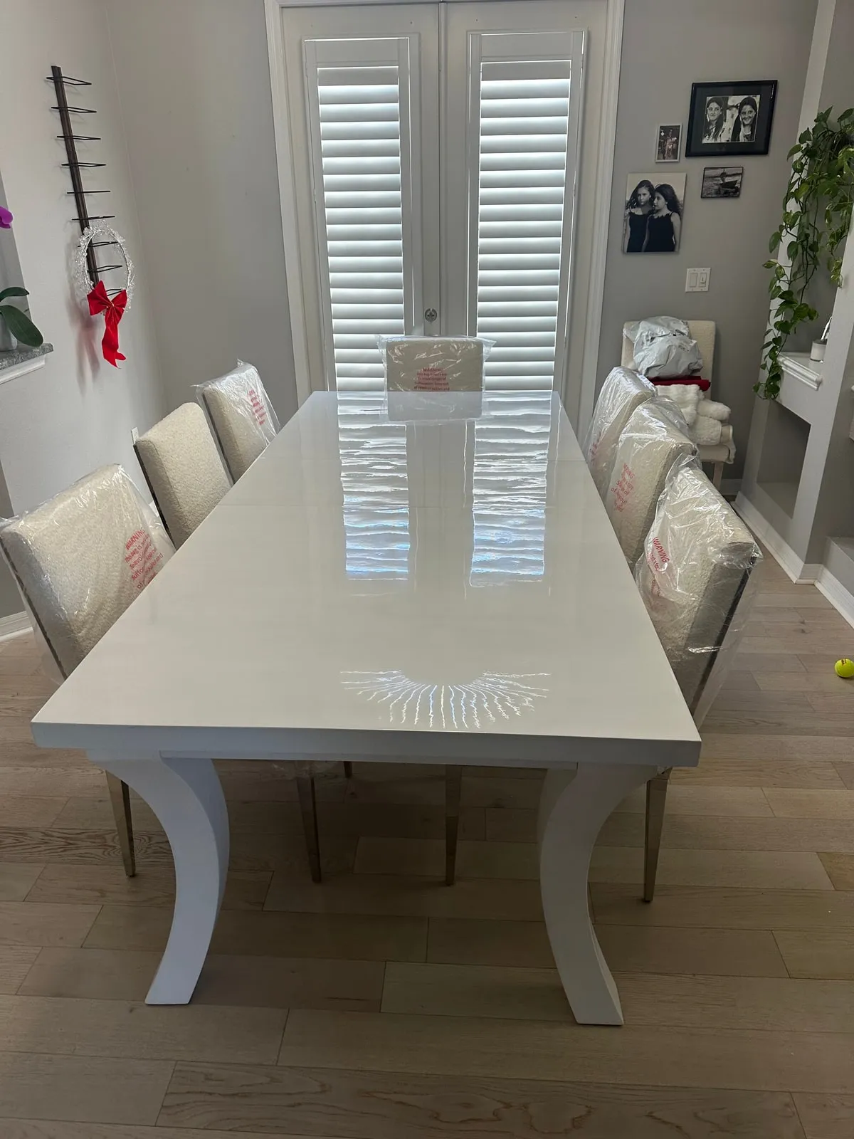 A rectangular dining table with a high-gloss off-white top and curved pale legs, photographed in a dining room where the upholstered chairs are still wrapped in factory plastic.